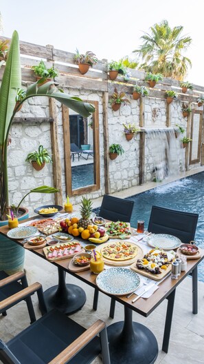 A dining table set with various dishes and drinks, surrounded by chairs, with a pool and plants in the background. - Ariel Hotel Alaçatı (Cesme)