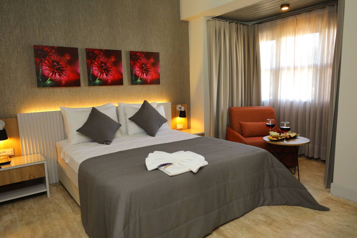 A hotel room with a bed, pillows, a robe, a chair, a table with a plate of food and glasses, and three wall-mounted flower paintings.