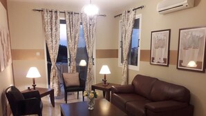 A living room with a brown leather sofa, two armchairs, a coffee table, and two lamps. There are two windows with curtains, an air conditioner, and two framed pictures on the wall. - Beautiful Apartment With Pool in Paphos, Cyprus (Paphos)