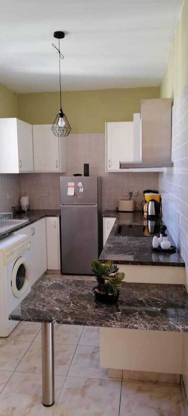 A kitchen with a stainless steel refrigerator, a washing machine, a marble countertop, and a hanging light fixture.