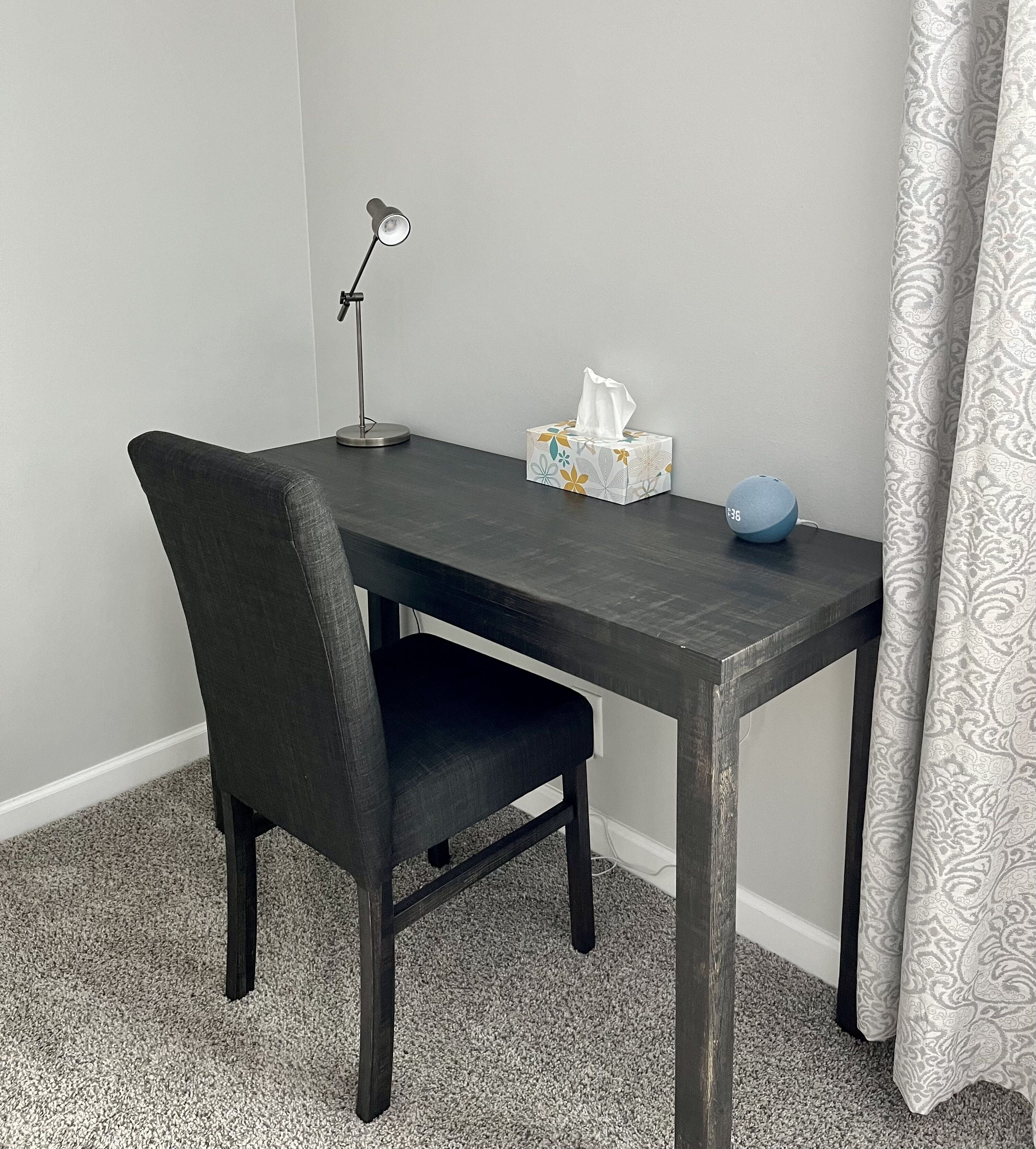 A small, dark wooden desk with a single chair, a desk lamp, a tissue box, and a blue speaker.
