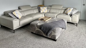 Un salon moderne avec un canapé d’angle beige, un pouf à motifs et une table basse dotée de télécommandes.