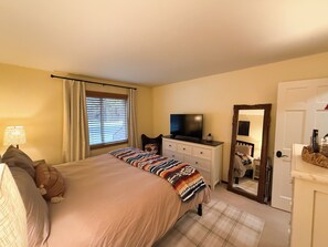 A bedroom with a bed, a television on a dresser, a window with blinds, and a mirror on the wall.