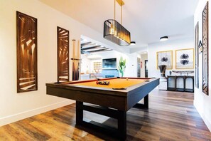 A pool table in a modern room with wooden flooring, a hanging light fixture, and framed artwork on the walls.