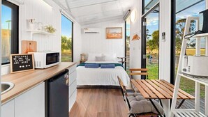 A compact room with a bed, a small kitchenette, a dining area, and large windows offering a view of the outdoors. - Avondale Heights by Tiny Away (Banyena)