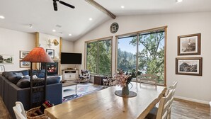 A spacious living room with a large wooden dining table, comfortable seating, a fireplace, and large windows offering a view of the outdoors. - Riverfront 4 bed/3bath home on Bitterroot River plus a Pickleball Court  (Hamilton)