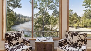 A room with two cow-print armchairs, a small wooden table with books, and a view of a river and forest through large windows. - Riverfront 4 bed/3bath home on Bitterroot River plus a Pickleball Court  (Hamilton)