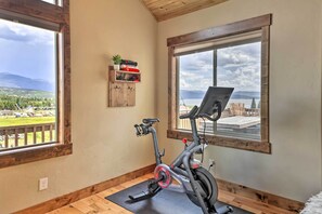 A room with a stationary bike, wooden flooring, a window with a view of a landscape, and a shelf with items. - Game Room & Hot Tub: Luxe Grand Lake Retreat! (Grand Lake)