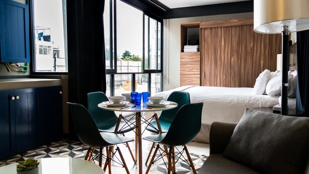 Moderni hotellihuone, jossa on suuri ikkuna, ruokailutila tuoleineen ja pöytineen, puinen päätyosaan varustettu sänky ja lamppu.
