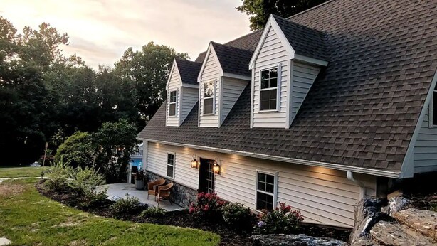 A two-story house with a sloped roof, a front porch with a bench, and a landscaped garden with rocks and shrubs.
