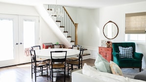 A spacious living room with a round dining table, black chairs, a green armchair, and a staircase.