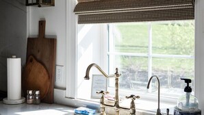 A kitchen with a white countertop, a stainless steel sink, a brass faucet, a wooden cutting board, a lantern, and a window with a brown roller blind.