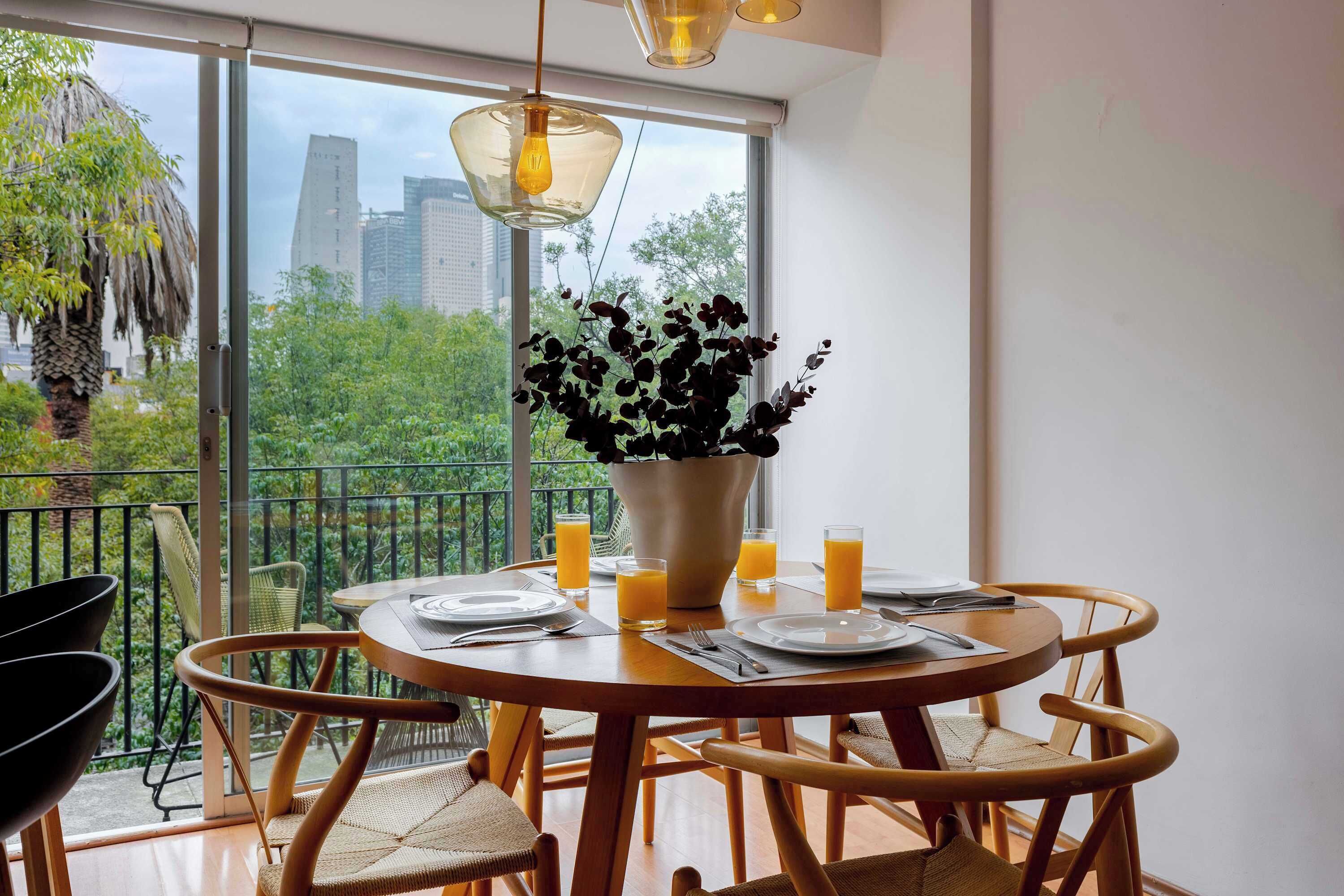 A dining area with a round table, chairs, a vase with flowers, and a city view through large windows.