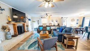 A spacious living room with a fireplace, a television mounted on the wall, and a dining area with a table and chairs. - Ground Level, Bayside Retreat w/ bay access! Dogs OK, Large Pond (Eastpoint)