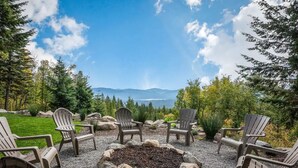 A fire pit surrounded by Adirondack chairs with a scenic view of mountains and a lake.