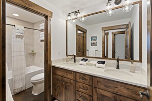 A bathroom with a bathtub, toilet, and a large mirror with lights. There are wooden cabinets and a white sink. - Gorgeous mountain getaway - VIEWS - HotTub - Private - Wildlife - Close to town (Sandpoint)