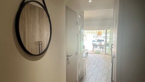 A hallway with wooden flooring, a circular mirror on the wall, and a door leading to a bright room with a view of the outside.