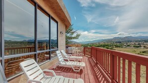 A balcony with white lounge chairs and a wooden deck, overlooking a scenic landscape with mountains and a clear sky.