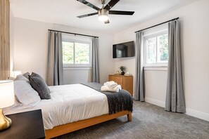 Une chambre à coucher avec un lit, un ventilateur de plafond, un téléviseur fixé au mur et une fenêtre avec des rideaux.