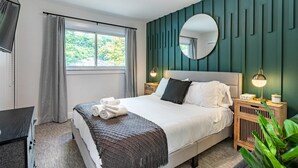 A neatly made bed with a white comforter and dark throw, a round mirror on the wall, and a bedside lamp.