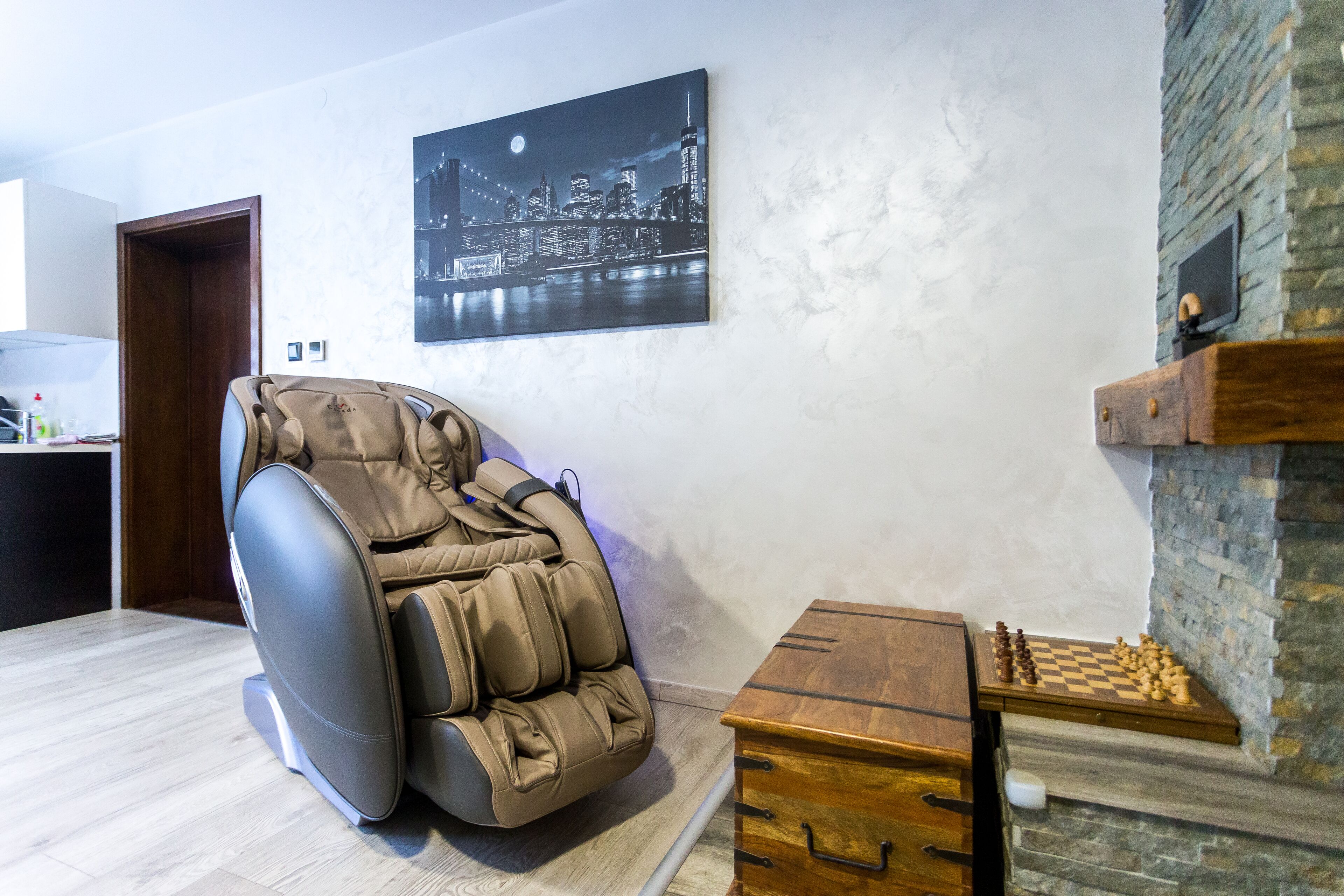 A massage chair, a chessboard, and a cityscape painting in a room with a wooden floor and stone wall.