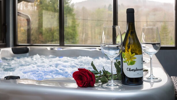 A hot tub with a bottle of Chardonnay and two glasses, a red rose, and a view of trees and mountains through large windows. - Holiday House Papuk is Located in Park of Nature (Brestovac)