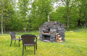 A stone-built outdoor fireplace with a chimney, surrounded by a grassy area with two wicker chairs.