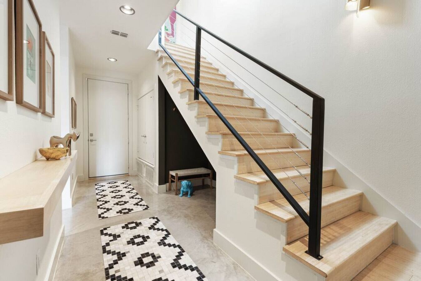 Una escalera con escalones de madera y barandilla de metal negro, que conduce a un piso superior. Una alfombra estampada cubre el suelo, y un pequeño banco está situado cerca de la escalera.