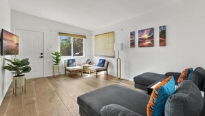 Ein modernes Wohnzimmer mit einem grauen Sofa, einem Couchtisch und einem Sessel. Zwei Fenster mit Vorhängen, eine Tür und ein an der Wand befestigtes Kunstwerk sind zu sehen.