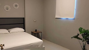 Un dormitorio con cama, una mesita de noche pequeña, una planta en maceta y una ventana con lámpara.