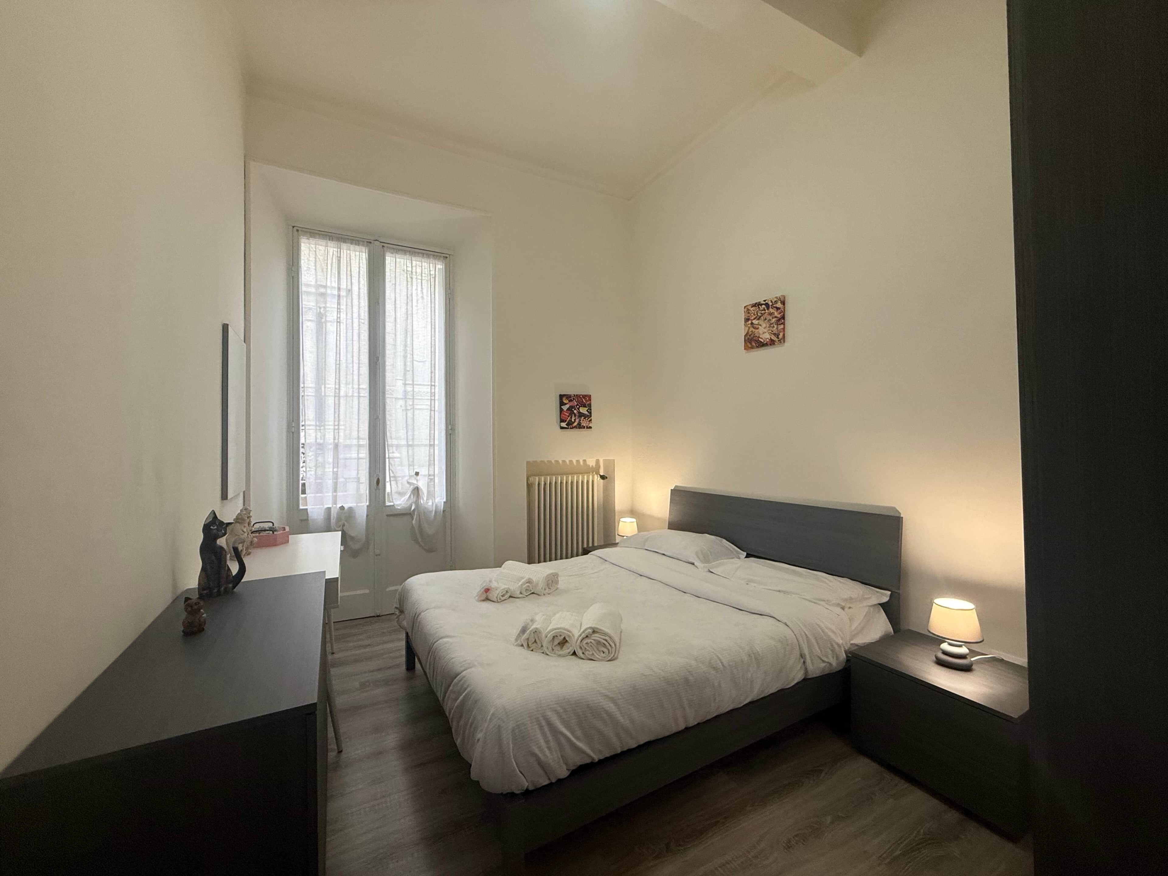 Un dormitori amb llit, taules de nit, un radiador i una finestra amb cortines.
