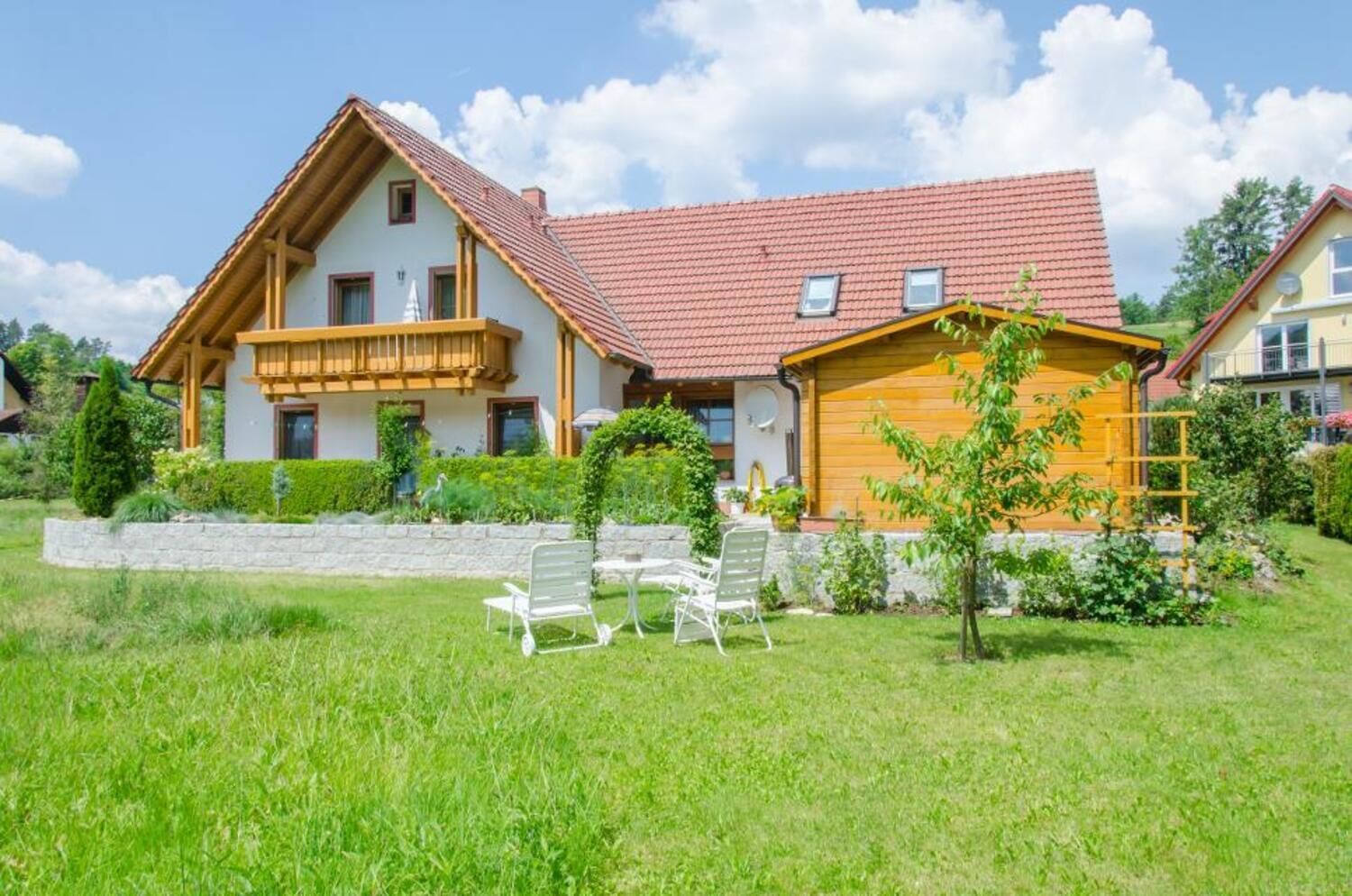 Ein zweistöckiges Haus mit rotem Ziegeldach, einem Holzbalkon und einem kleinen Garten mit zwei weißen Stühlen.