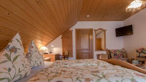 A bedroom with a wooden ceiling, a bed with floral bedding, a desk with a lamp, a chair, a dresser, a mirror, and a television on the wall.