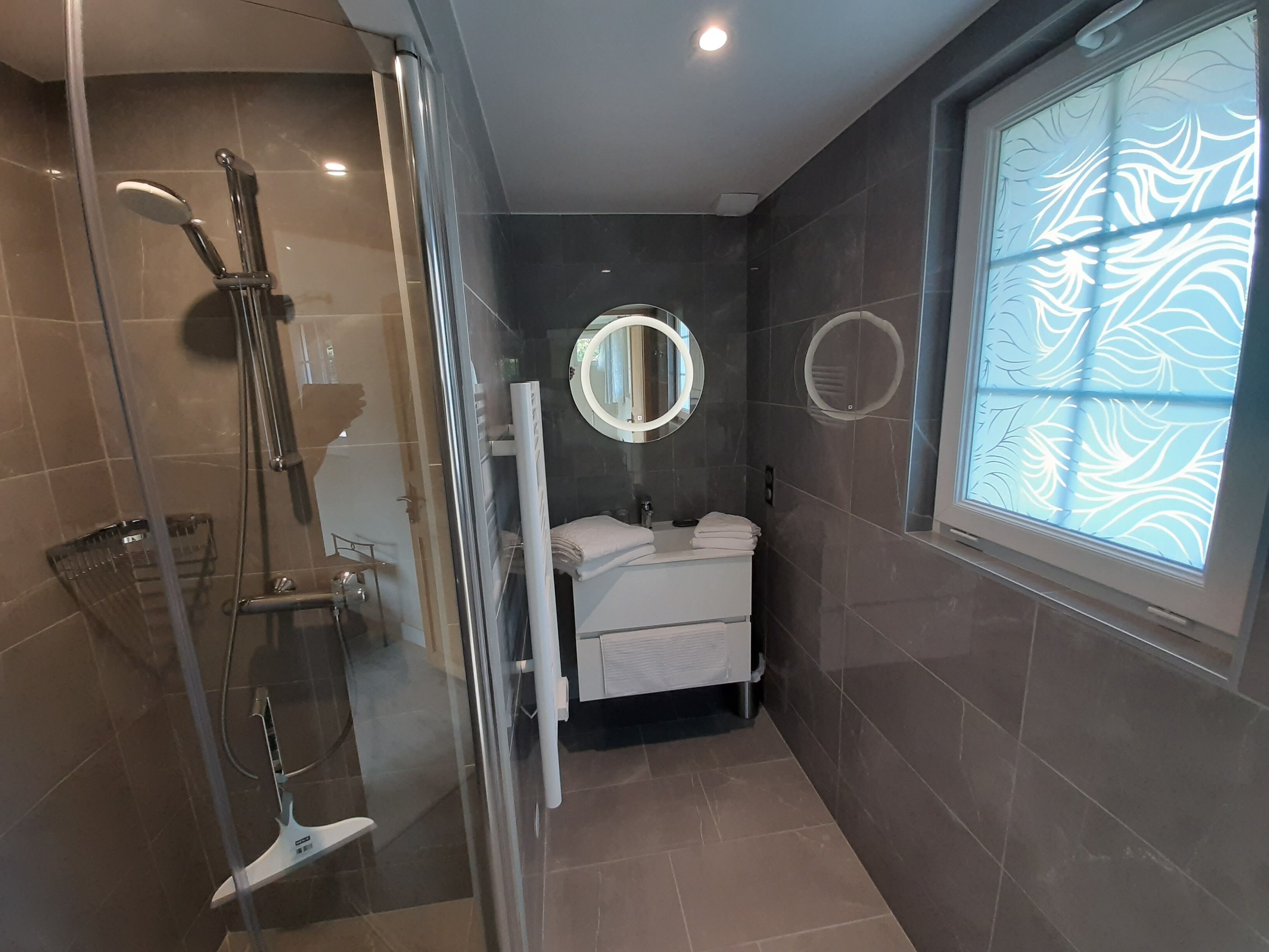 Une salle de bain moderne avec une douche à parois de verre, un miroir rond et une fenêtre ornée d’un motif de feuilles.