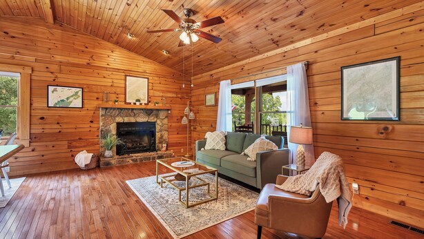 A wooden cabin interior with a fireplace, a green sofa, a wooden ceiling fan, and a dining area with a table and chairs. - This Beautiful Cabin with Mountain views is the Perfect getaway! (Townsend)