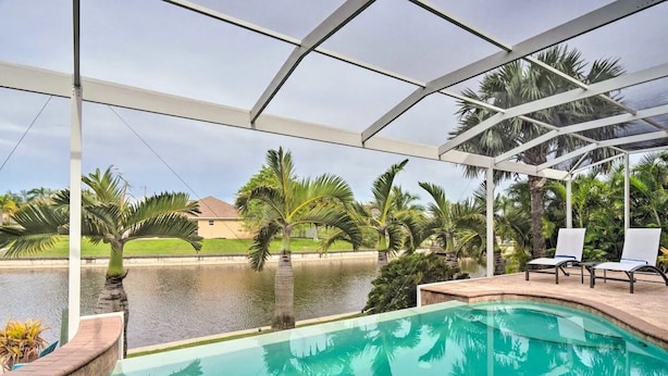 Una piscina al aire libre con cerramiento de vidrio, palmeras y vista a una casa al otro lado del agua.