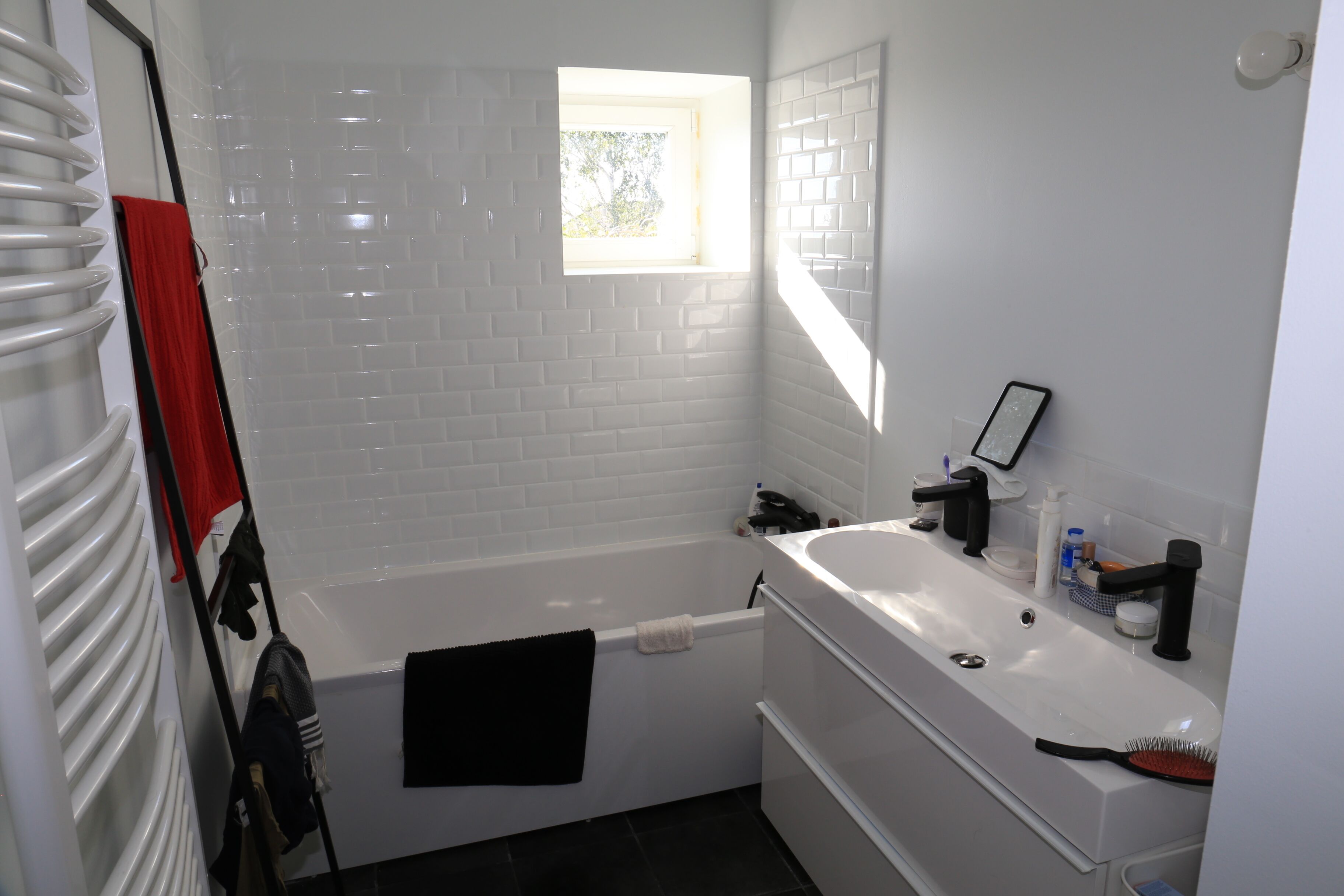 A bathroom with a white bathtub, a window, a mirror, and a sink with toiletries.