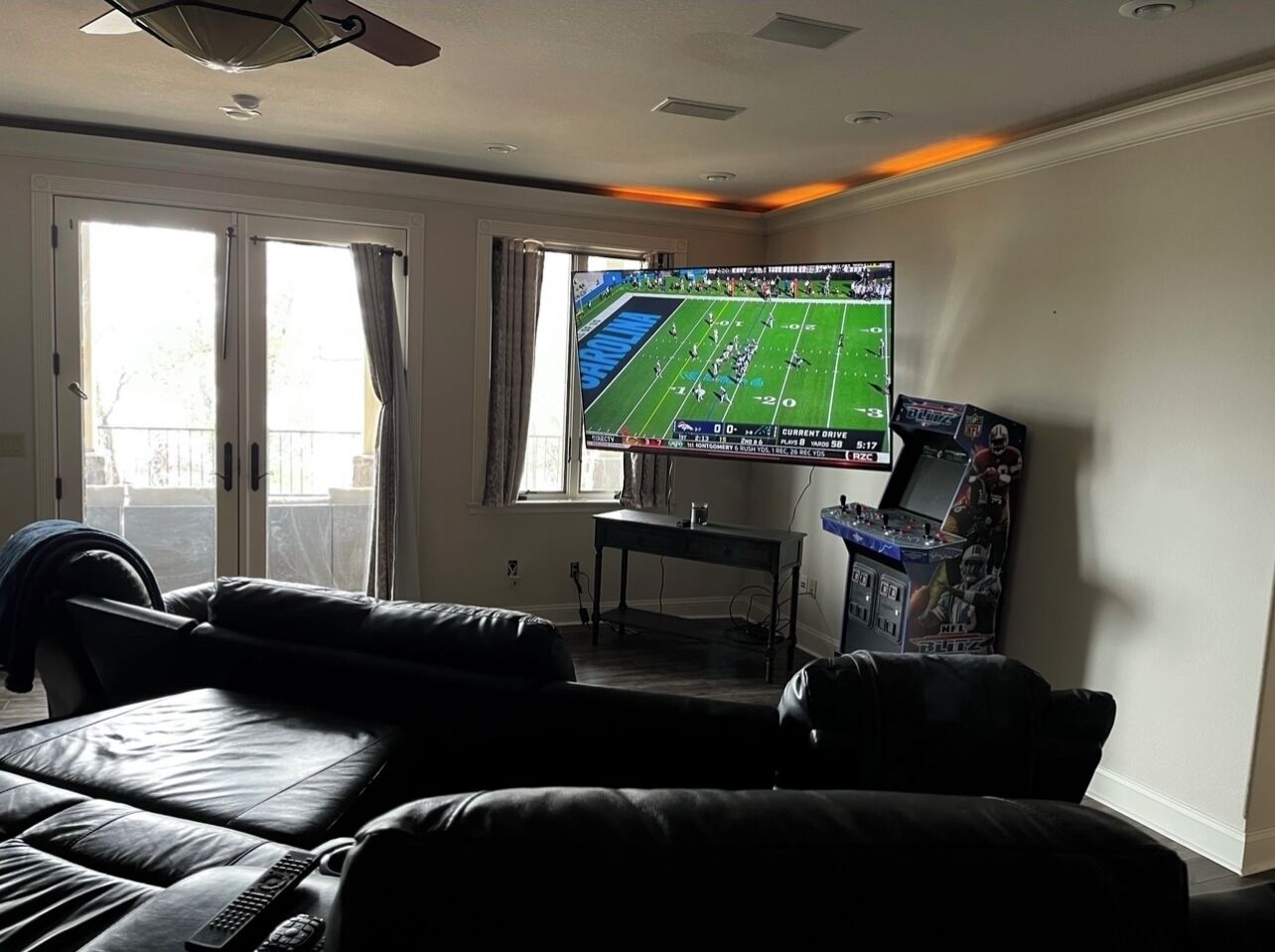 Ein Wohnzimmer mit einem großen Flachbildfernseher, der ein Fußballspiel zeigt, einer Ledersofa, einem kleinen Tisch und einer Spielkonsole.