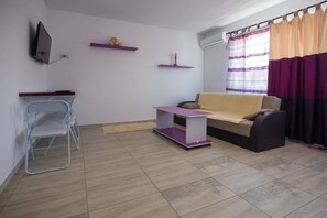 Un salon avec un canapé, une petite table, un téléviseur fixé au mur et une fenêtre avec des rideaux.