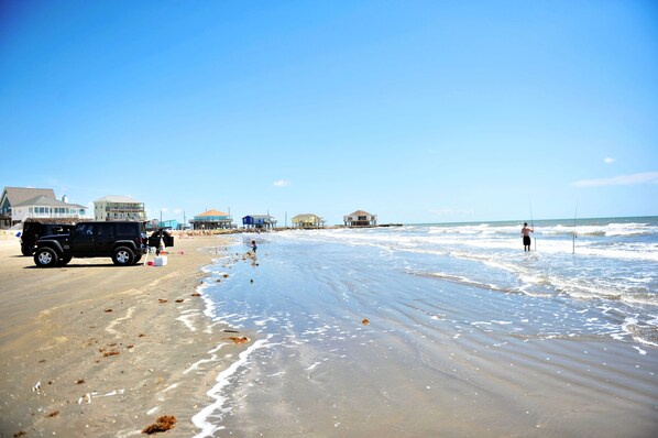 A beach with a black vehicle parked on the sand, a person fishing, and several buildings in the background.