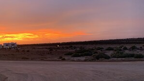 Een woestijnlandschap met een zonsondergang, enkele gebouwen en schaarse vegetatie.