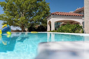 A swimming pool with clear blue water, surrounded by greenery and a building with a tiled roof.