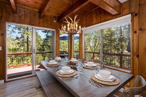 A wooden dining area with a large table set for six, large windows, and a chandelier. - New! Rustic & cozy cabin w/pool table, deck, garage, dog friendly! (Lake Arrowhead)