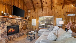 Ein gemütliches Wohnzimmer mit Kamin aus Naturstein, Flachbildfernseher, großem Fenster und bequemen Sofa.
