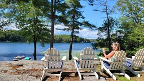 A lakeside view with four Adirondack chairs facing the water, a boat docked nearby, and a clear sky.