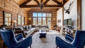 A log cabin interior with a large sofa, two blue armchairs, a coffee table, and a mounted deer head on the wall.
