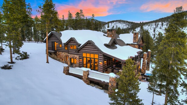 A log cabin with a stone chimney, large windows, and a snow-covered landscape.