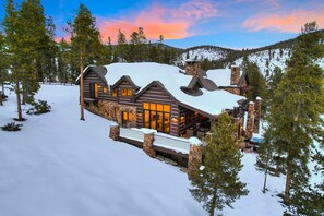 A log cabin with a stone chimney, large windows, and a snow-covered landscape.