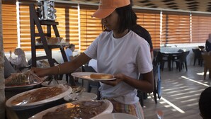A person serving food at a buffet table with various dishes and a food display stand in a dining area with wooden blinds and tables. - Alena Hotel (Aqaba)