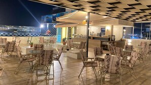 An outdoor seating area with tables and chairs, a bar area with a counter, and a city view through large windows. - Alena Hotel (Aqaba)